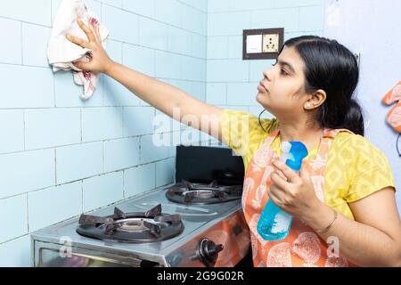 Eine hübsche indische junge Frau, die Schürze trägt und die Küche mit Küchenreiniger putzt Stockfoto