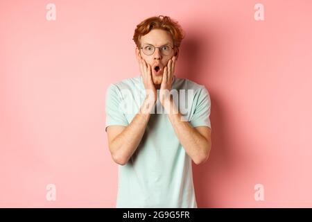 Schockiert Rotschopf Mann Klatschen, starrte beeindruckt auf Kamera und berühren Gesicht, stehend in einer Brille vor rosa Hintergrund Stockfoto