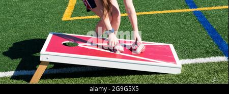 Horizontale Ansicht eines Mädchens, das gestreifte Bohnenbeutel aufnimmt, während es auf einem selbstgemachten rot-weißen Brett Cornhole spielt. Stockfoto