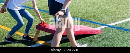 Seitenansicht von zwei Mädchen, die mit einem roten selbstgemachten Brett auf einem Rasenfeld ein Cornhole spielen und Bohnenbeutel abheben und werfen. Stockfoto