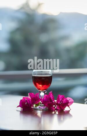 Ein Glas Rotwein auf unscharfem Berghintergrund. Dekorationen von rosa Bougainvillea Blumen. Blue Hour Fotografie. Speicherplatz kopieren. Stockfoto