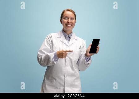 Porträt einer Ärztin, therapeutischen oder medizinischen Fachberaterin mit einem isolierten Smartphone-Bildschirm auf blauem Hintergrund. Lustige glückliche Emotionen. Konzept von Stockfoto