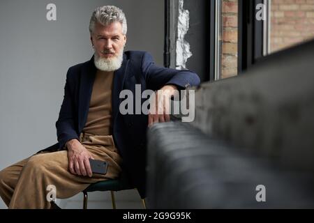 Stilvoller grauhaariger Mann mittleren Alters in legerer Kleidung, der die Kamera anschaut, das Smartphone hält, während er an einem Fenster im modernen Loft-Interieur sitzt Stockfoto