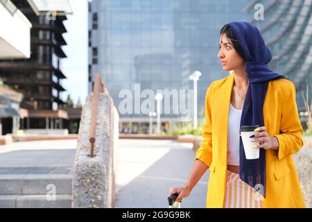 Junge indische Geschäftsfrau in gelber Anzugjacke beim Spazierengehen und mit einer Tasse Kaffee und einem Koffer im Freien. Stockfoto