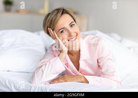 Portrait einer schönen blonden Frau mittleren Alters mit kurzem Haarschnitt in pinker Seide, die zu Hause auf den weißen Bettlaken liegt, süßer weiblicher Anschauung Stockfoto