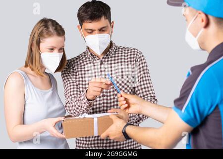 Das junge Paar in medizinischen Masken Mann und Frau haben das Paket vom Lieferer erhalten und gibt dem Mann Stift, um dafür zu unterschreiben. Stockfoto