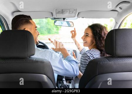 Fröhlicher junger arabischer Mann und Frau in legerem Outfit, der Spaß hat, während er mit dem Auto in den Urlaub fährt, tanzt und Lieder singt, aufgenommen vom Auto-Rücksitz. Unterhaltung Stockfoto
