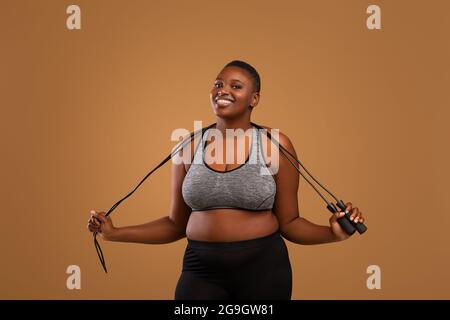 Fitness Plus Size Modell. Porträt Einer Selbstbewussten, Lächelnden Jungen Afroamerikanischen Frau In Sportswear Holding Springseil Auf Den Schultern, Posiert Beim Brown Stud Stockfoto