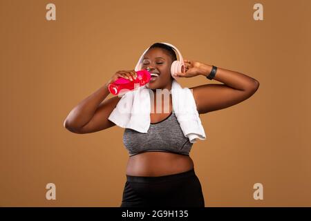 Sportversorgung. Porträt einer kurvigen afroamerikanischen Frau mit weißem Handtuch am Hals, das frisches Wasser aus der Flasche trinkt, kabellose Kopfhörer trägt und Stockfoto