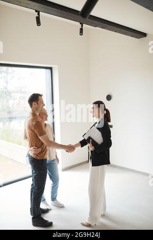 Vertikale Seitenansicht Porträt eines jungen Paares, das sich während der Tour durch die Wohnung mit dem Immobilienmakler die Hände schüttelt, Platz kopieren Stockfoto