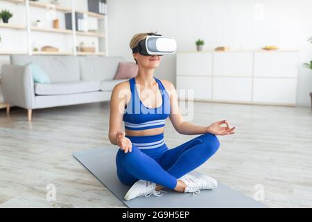 Moderne Technologien Und Sport. Ruhige, friedliche Frau mit VR-Headset, die in Lotushaltung sitzt, zuhause meditiert und Yoga praktiziert. Dame usi Stockfoto