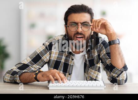 Verwirrt Mann aus dem Nahen Osten Programmierer sitzen am Arbeitstisch, mit modernen Miniatur-pc-Tastatur, berühren Brille, Nahaufnahme Porträt. Arabischer Mann, Freiberufler Stockfoto