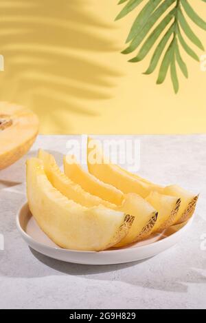 Weißer Teller mit reifen Melonenscheiben auf gelbem Hintergrund mit Palmenblatt-Schatten, Sommerfrüchten. Stockfoto