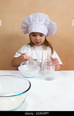 Ein kleines Mädchen, das als Köchin gekleidet ist, knetet den Teig. Kochen Kind Lifestyle-Konzept. Das Kind liebt, hat Spaß, studiert und spielt in der Küche Stockfoto