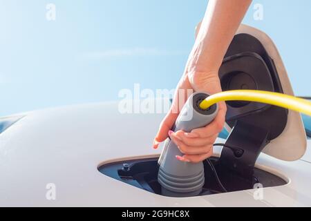 Weibliche Hand, öffnen Sie eine Steckdosenabdeckung und stecken Sie das Ladegerät in ein Elektroauto an einer Ladestation Stockfoto