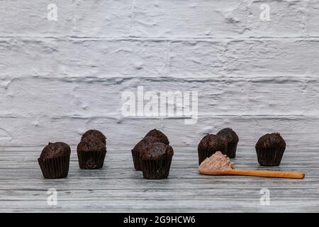 Auf Holzbrettern befinden sich mehrere kleine Schokoladen-Cupcakes und ein Holzlöffel mit Schokoladenpulver und im Hintergrund eine weiße Ziegelwand Stockfoto