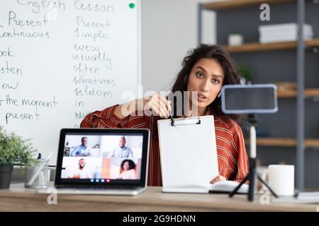 Online-Präsentation. Porträt einer jungen Tutorin in Ohrhörern, die auf ein leeres Blatt Papier zeigt und auf ein Mobiltelefon auf einem Stativ zeigt. Frau mit vir Stockfoto