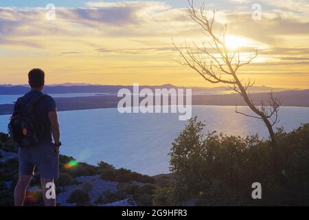 Männlicher Wanderer auf dem Hügel, der einen wunderschönen Sonnenuntergang über der Adria beobachtet. Konzepte für Wandern, Leistung, Erwartung, Optimismus und Selbstreflexion. Stockfoto