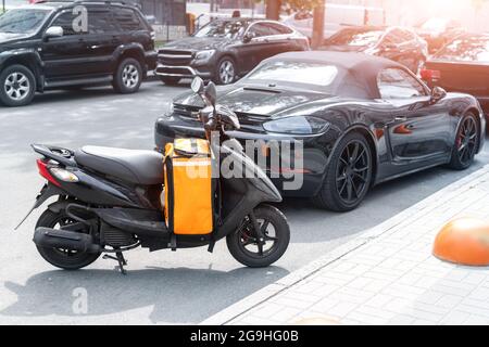 Schnelle Express Takeaway Online-Gig bestellen Lebensmittel Lieferung Roller mit rot orange Tasche Box geparkt in Verkehr Stadtzentrum Innenstadt Straße an sonnigen Tag. Essen Stockfoto