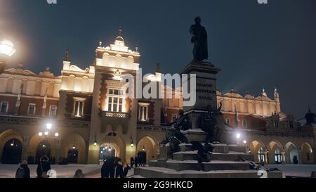 Krakau, Polen 03.02.2021 der Tuchsaal (oder die Sukiennice) mitten in der Krakauer Altstadt. Die Statue von Adam Mickiewicz in der Nacht vor dem Tuchsaal. Touristen, die auf der Straße herumlaufen. Stockfoto