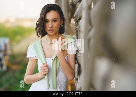 Asiatische Frau, die in der Nähe eines Tabaktrocknungsschuppens posiert und ein weißes Kleid und grüne Gummistiefel trägt. Stockfoto