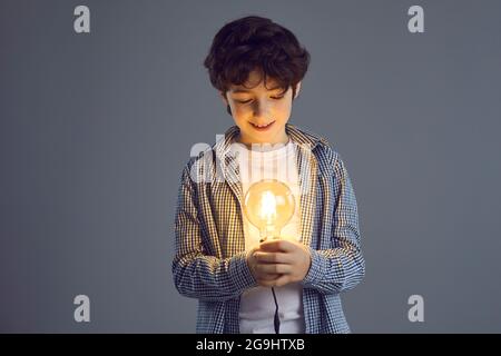 Kleiner lächelnder lockiger Junge steht auf einem grauen Hintergrund und blickt auf eine helle Glühbirne in seinen Händen. Stockfoto