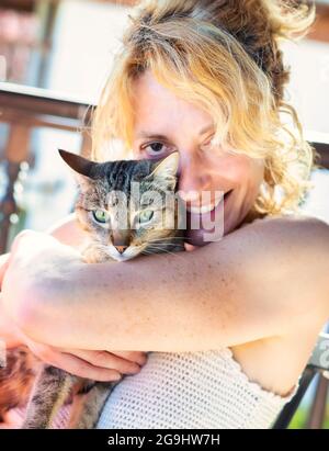 Porträt einer jungen reifen kaukasischen blonden Frau, die mit ihrer Katze zu Hause auf der Veranda lächelt und in einem Schaukelstuhl sitzt. Lifestyle-Konzept. Stockfoto