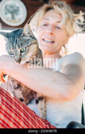 Porträt einer jungen reifen kaukasischen blonden Frau, die mit ihrer Katze zu Hause auf der Veranda lächelt und in einem Schaukelstuhl sitzt. Lifestyle-Konzept. Stockfoto