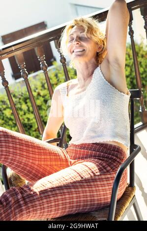 Porträt einer jungen reifen kaukasischen blonden Frau, die sich zu Hause auf der Veranda entspannt und in einem Schaukelstuhl sitzt. Lifestyle-Konzept. Stockfoto