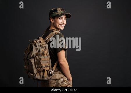 Selbstbewusst lächelnde Soldatin posiert mit Rucksack isoliert über der schwarzen Wand Stockfoto