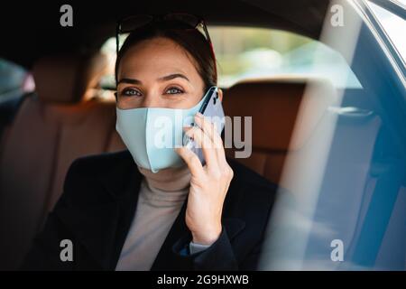 Bild einer erstaunlichen Geschäftsfrau, die in einem Auto in Gesichtsmaske sitzt und mit dem Mobiltelefon spricht Stockfoto