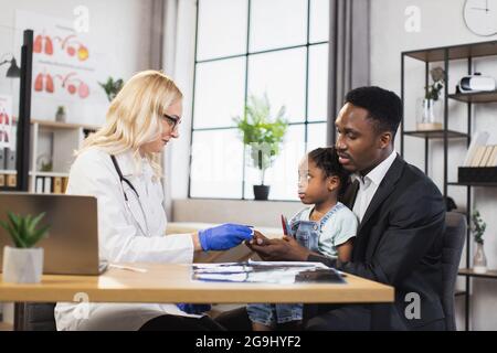 Kaukasischer Arzt, der die Sauerstoffsättigung eines kleinen afrikanischen Mädchens mithilfe eines modernen Pulsoximeters überprüft. Nette Patientin sitzt auf den Knien ihres fürsorglichen Vaters. Fa Stockfoto