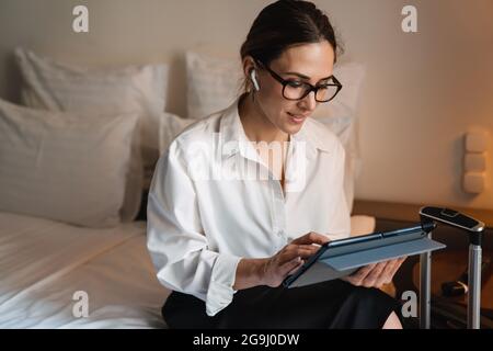 Eine brünette, weiße Geschäftsfrau mittleren Alters, die in einem Hotelzimmer auf dem Bett sitzt und das Tablet mit kabellosen Kopfhörern anfasst Stockfoto