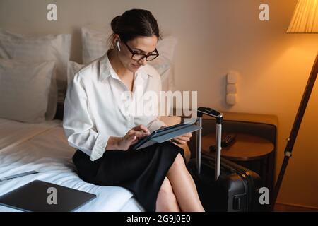 Eine brünette, weiße Geschäftsfrau mittleren Alters, die in einem Hotelzimmer auf dem Bett sitzt und das Tablet mit kabellosen Kopfhörern anfasst Stockfoto
