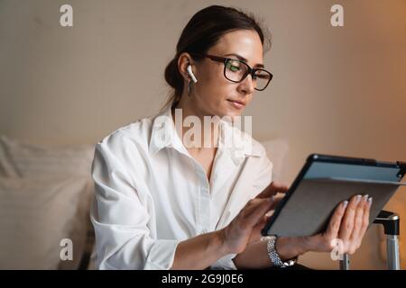 Eine brünette, weiße Geschäftsfrau mittleren Alters, die in einem Hotelzimmer auf dem Bett sitzt und das Tablet mit kabellosen Kopfhörern anfasst Stockfoto