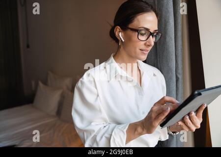 Lächelnde, brünett-weiße Geschäftsfrau mittleren Alters in smarter Kleidung, die am Fenster im Hotelzimmer mit kabellosen Ohrhörern steht und Tablet-Entwickler anfasst Stockfoto
