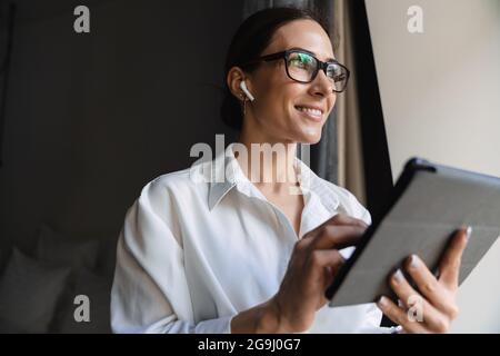 Lächelnde, brünett-weiße Geschäftsfrau mittleren Alters in smarter Kleidung, die am Fenster im Hotelzimmer mit kabellosen Ohrhörern steht und Tablet-Entwickler anfasst Stockfoto