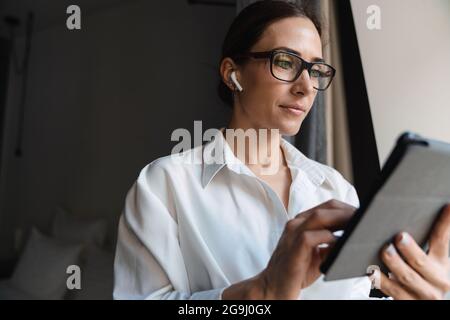 Lächelnde, brünett-weiße Geschäftsfrau mittleren Alters in smarter Kleidung, die am Fenster im Hotelzimmer mit kabellosen Ohrhörern steht und Tablet-Entwickler anfasst Stockfoto