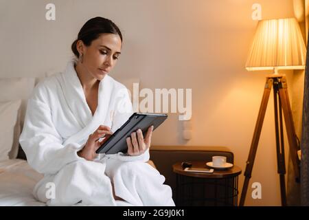 Lächelnde Brünette im mittleren Alter weiße Frau im Bademantel sitzt auf dem Bett drinnen Stockfoto