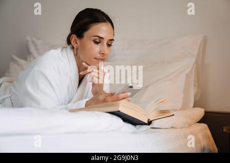 Lächelnde, brünette weiße Frau mittleren Alters im Bademantel, die drinnen auf dem Bett liegt und ein Buch liest Stockfoto