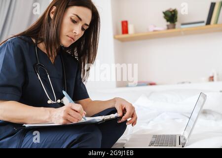 Eine Seitenansicht einer Brünette in einem medizinischen Anzug mit Stethoskop, die vor einem Laptop sitzt und etwas mit Papieren auf einem Tablet schreibt Stockfoto