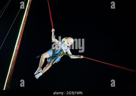 Kleines Mädchen mit blonden Haaren in der Luft auf dem Katapult reiten gegen den schwarzen Himmel. Lustige Sommerferien. Stockfoto