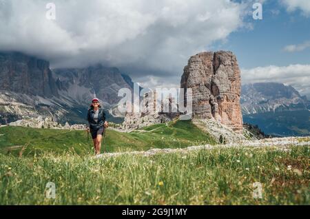 Lächelnde weibliche Trekkinginnen wandern mit Rucksack und Trekkingstöcken am grünen Berghang mit malerischer Dolomitenalpen Cinque Torri Formation auf der BA Stockfoto