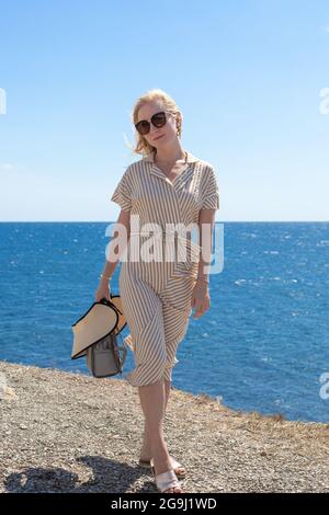 Eine schöne blonde Frau in einem Kleid läuft entlang der Küste. Sommerurlaub, Reisen und Tourismus. Entspannen an einem sonnigen Tag. Stockfoto