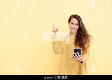 Lächelnde junge Frau hält ein Handy mit leerem Bildschirm und zeigt ihren Zeigefinger zur Seite. Stockfoto