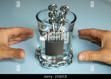 Ein Glas klares Wasser, in eine Kette gewickelt und mit einem Vorhängeschloss verschlossen, die Hände nach außen gestreckt, Wasserproblem, vermindernde Ressourcen von frischem Wat Stockfoto
