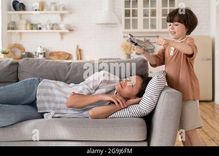 Gestresste Mutter versuchen, auf dem Sofa zu schlafen leiden unter Kopfschmerzen von frechen Sohn bang Metall Küchengeschirr Stockfoto