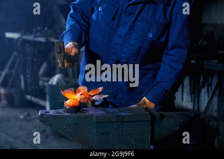 Professioneller Schmied in Uniform mit schwerem Hammer zum Schmieden von geschmolzenem Metall. Nahaufnahme der männlichen Hände mit Schnur. Arbeiten Sie bei FORGE. Stockfoto