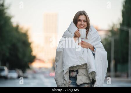 Bokeh-Porträt einer Frau, die in eine Decke gehüllt ist und Kaffee über dem Hintergrund der Stadt trinkt. In den frühen Morgenstunden. Stockfoto