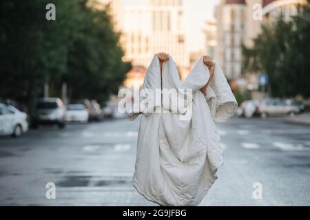 Person in Decke in der Mitte der leeren nassen Stadtstraße am frühen Morgen gewickelt. Ghost wird gespielt. Stockfoto